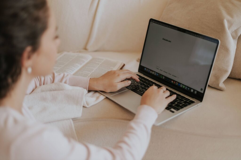 pexels-photo-35963245-35963245 Adult woman typing on a laptop with an open book nearby, working comfortably from her sofa.